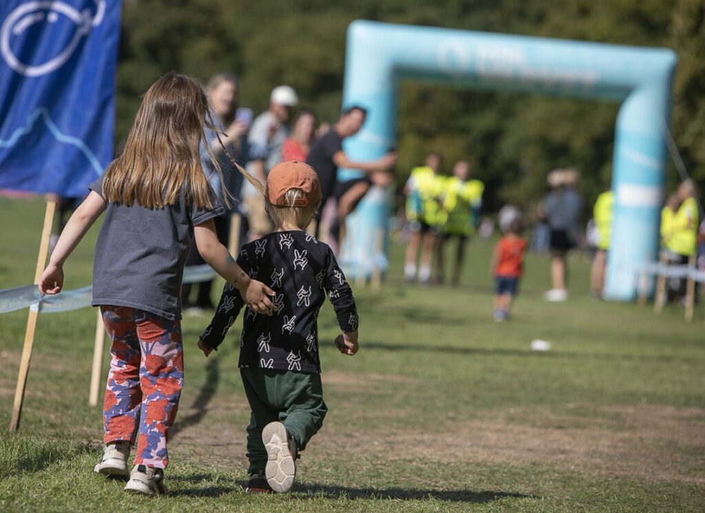 två barn springer ett lopp på en gräsmatta mot målet. i bakgrunden syns vuxna personer klappa händer och heja på
