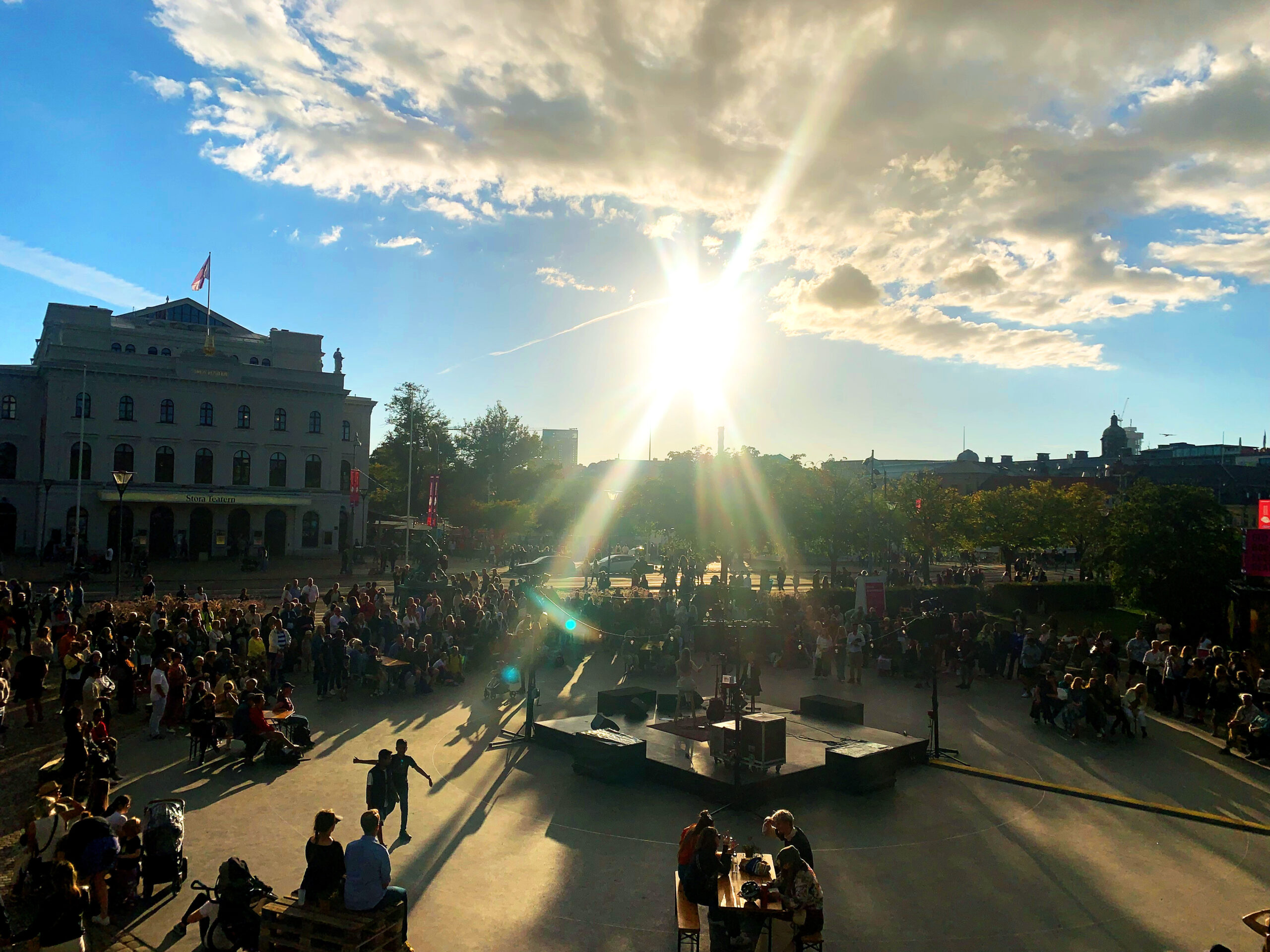 In the middle of a park there is an open stage. Around the stage people are waiting for the next performer. Sun is setting and makes a warm light. In the left of the photo theres a large, old building known as Stora Teatern.
