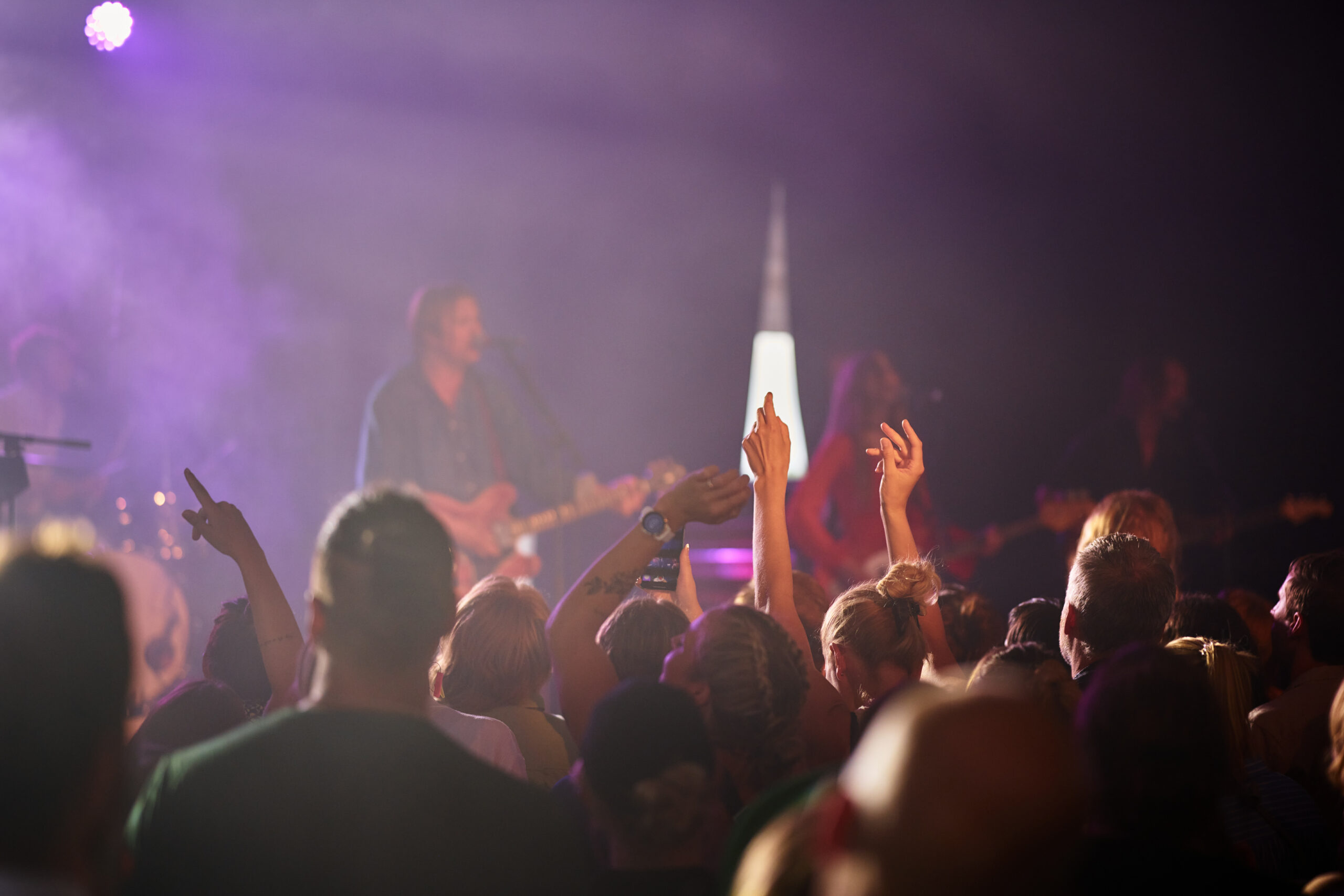 publik som trängs framför en scen där ett band spelar. fokus ligger på en persons händer som är uppsträckta i luften