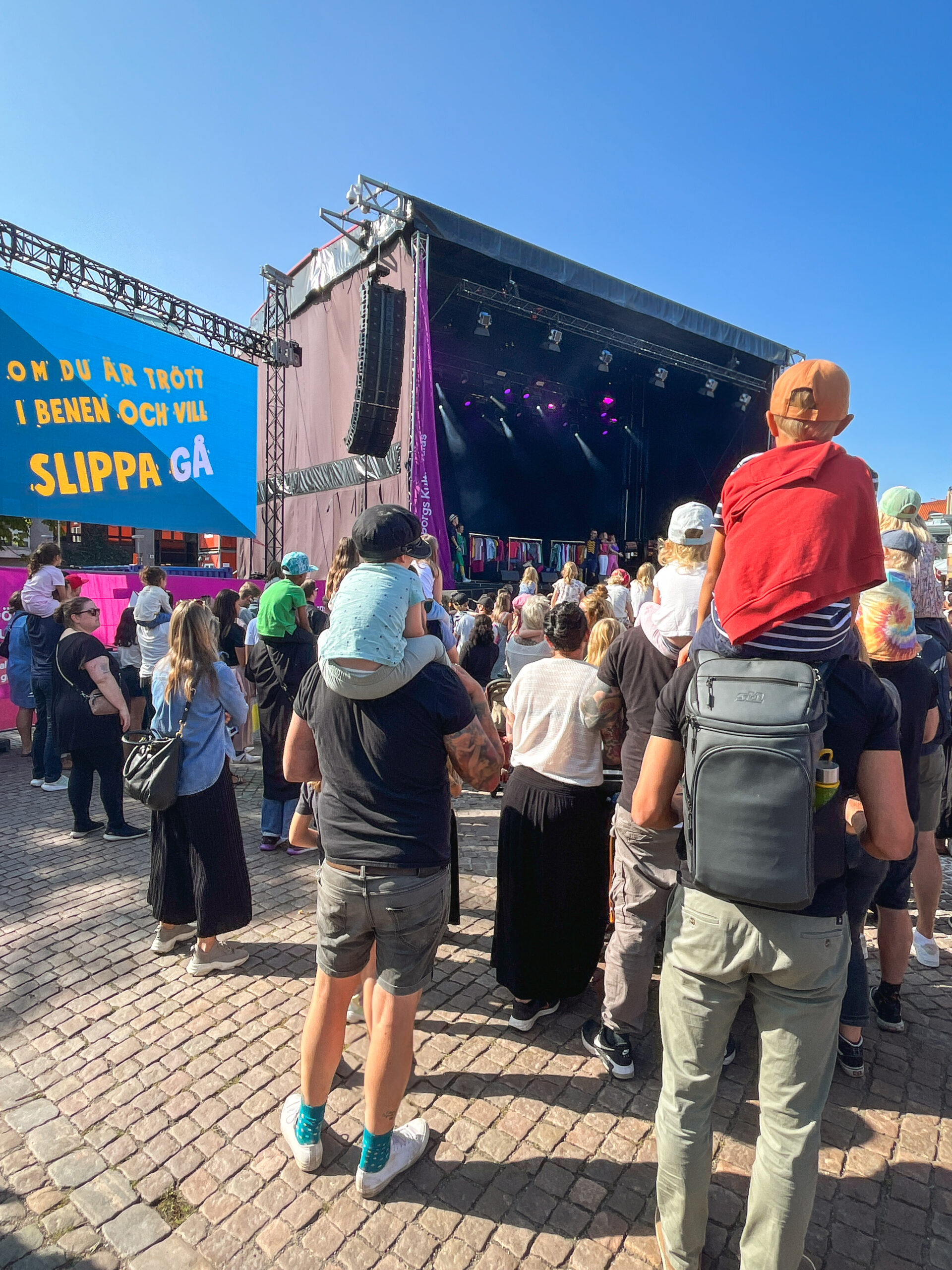 Kokobäng performs on a large stage. The camera's gaze is on children sitting on adults' shoulders with their backs to the camera. On the left in the picture you can see a large screen with the text "om du är trött i benen och vill slippa gå" (eng: "if you are tired in the legs and don't want to walk") which is the text from the song that Kokobäng sings. It is a clear day with blue skies.
