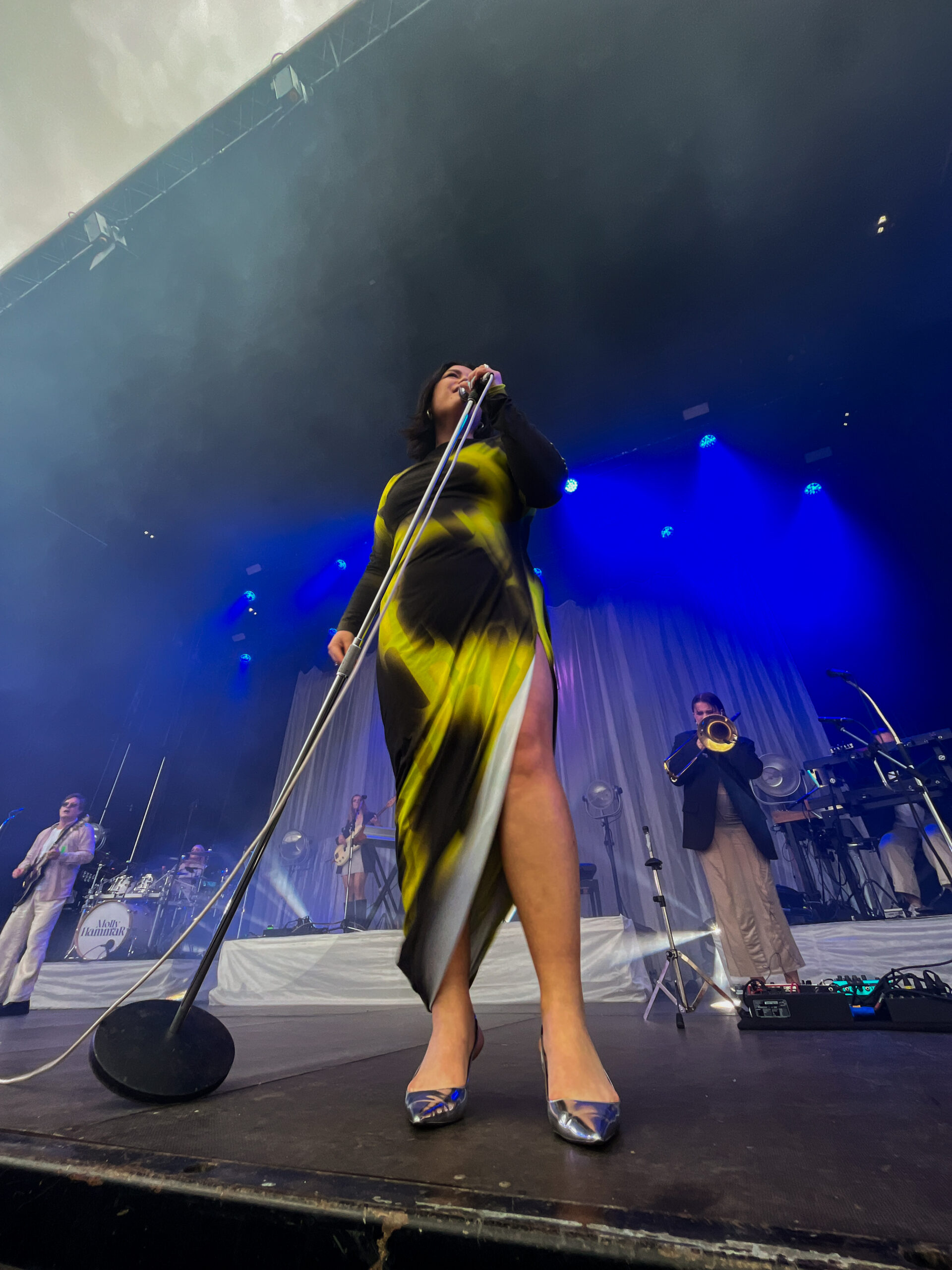 Molly Hammar performs on stage in a black and yellow long dress. She is holding a mic stand and singing. The band and the big stage can be seen in the background.