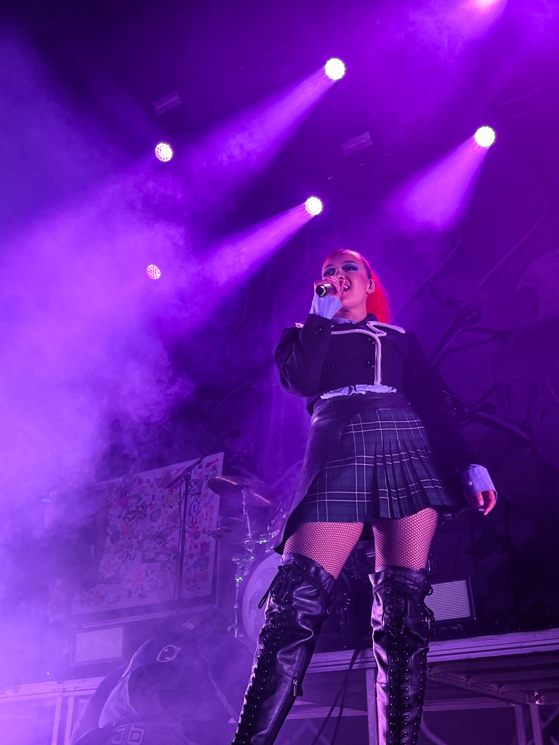 Daniela Rathana sings on stage. She wears tall leather boots, a short plaid skirt and short black blazer. Her hair is bright red and put up in a high ponytail. A drum set is visible in the background