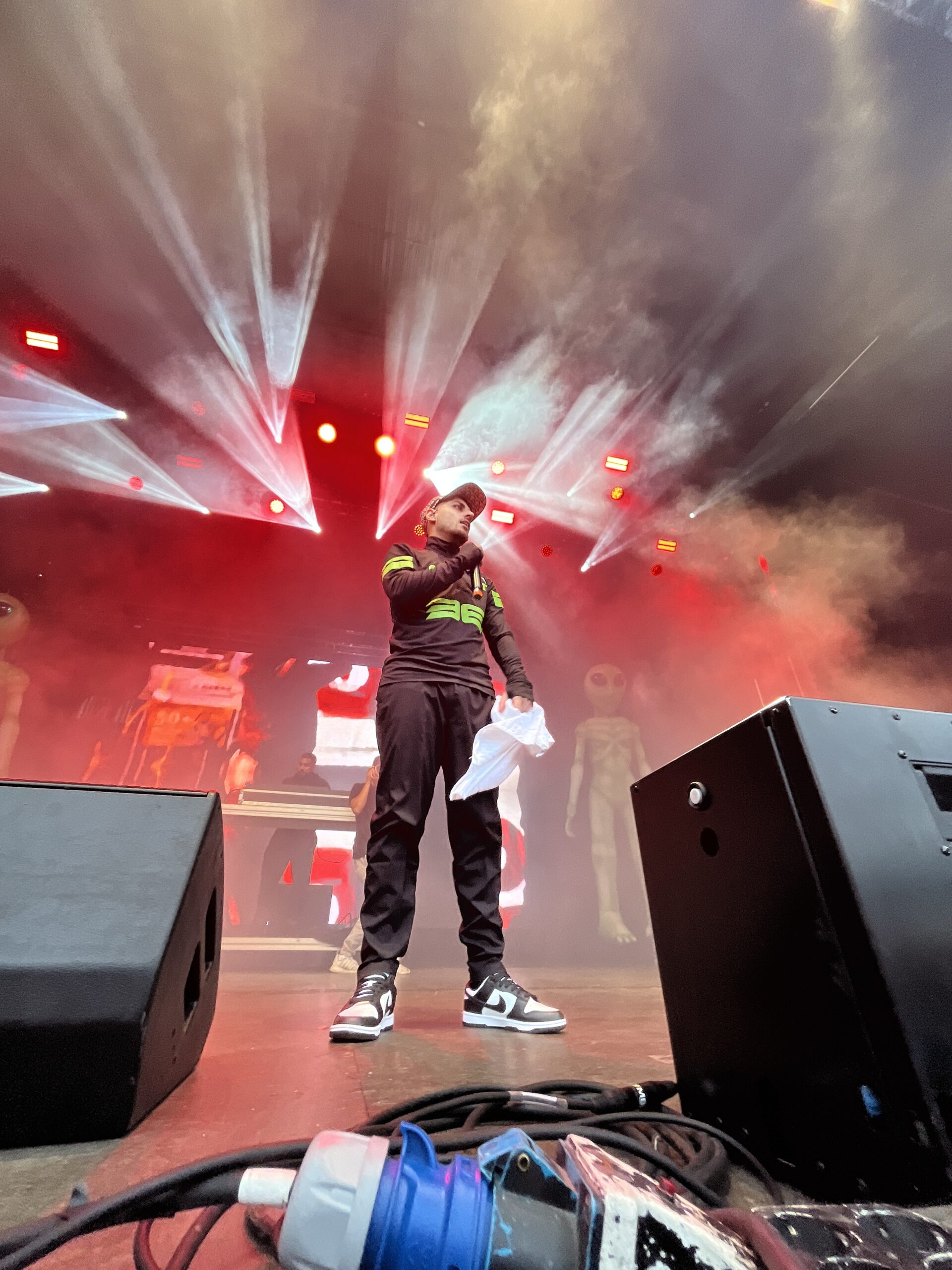 A36 performs on stage wearing tracksuit, Nike shoes and a cap. He holds a white towel in one hand and a microphone in the other. Red and white lights shine in the background.