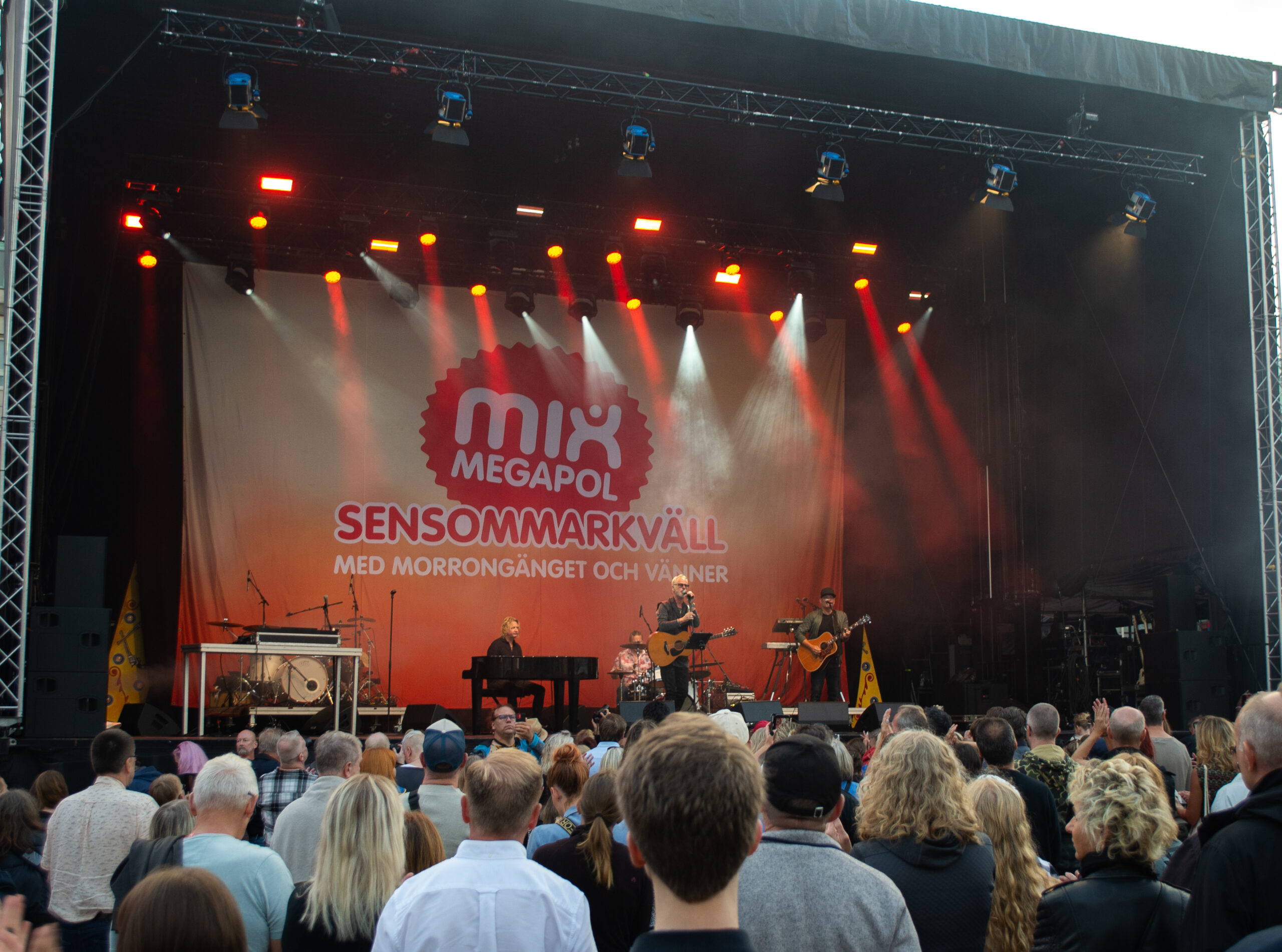 Uno Svenningsson plays on a stage with a band. In the background of the stage hangs a sign that says Mix Megapol Sensommarkväll (eng: Late Summer Night). In the foreground, the audience can be seen enjoying the concert.