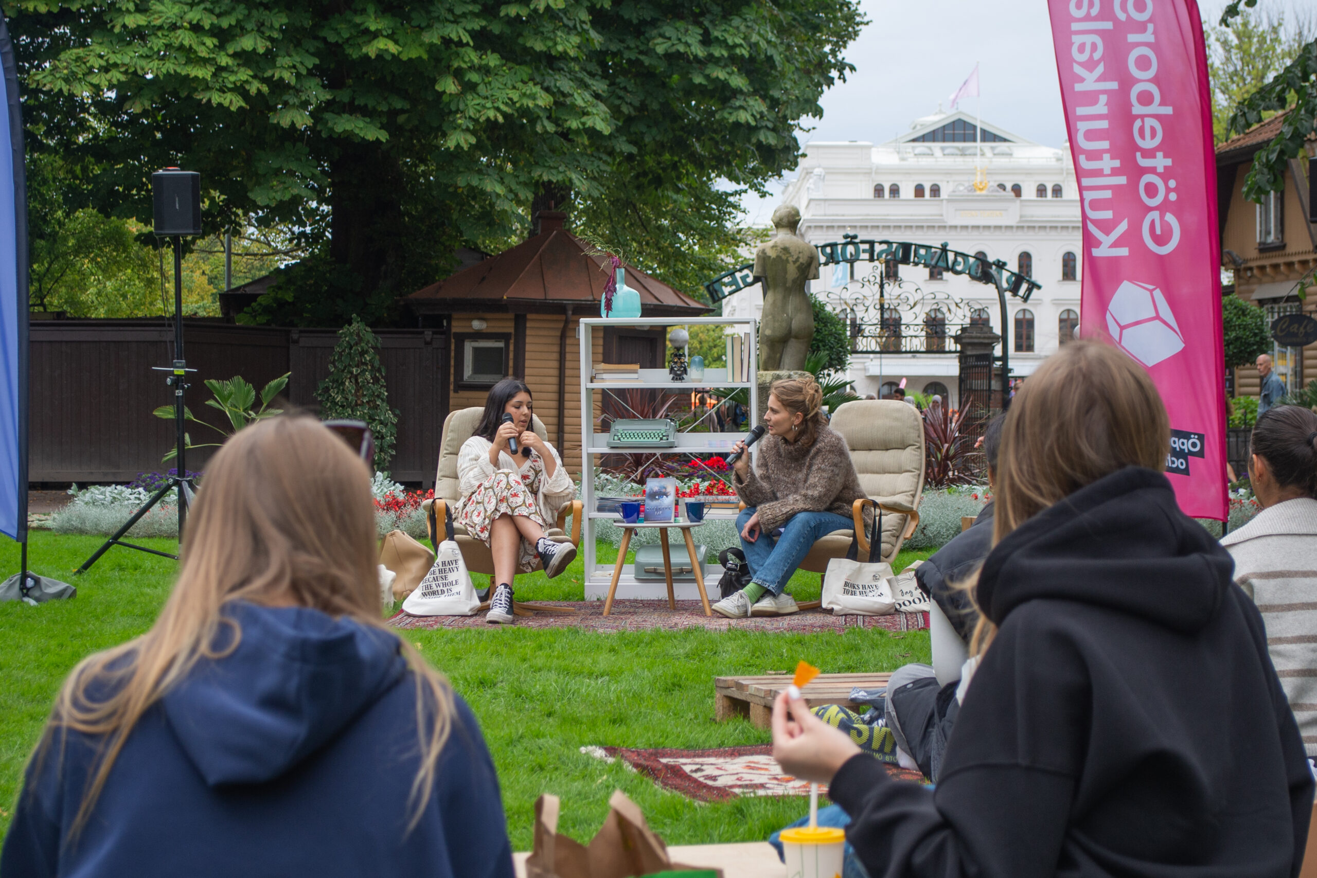 Jasmine Darban and a person from Bokmässan (eng: the Book Fair) discuss books. They each sit in a beige and cozy armchair in the Garden Association. In the foreground, people are seen listening and having coffee, they have their backs to the camera and are sitting on a blanket.