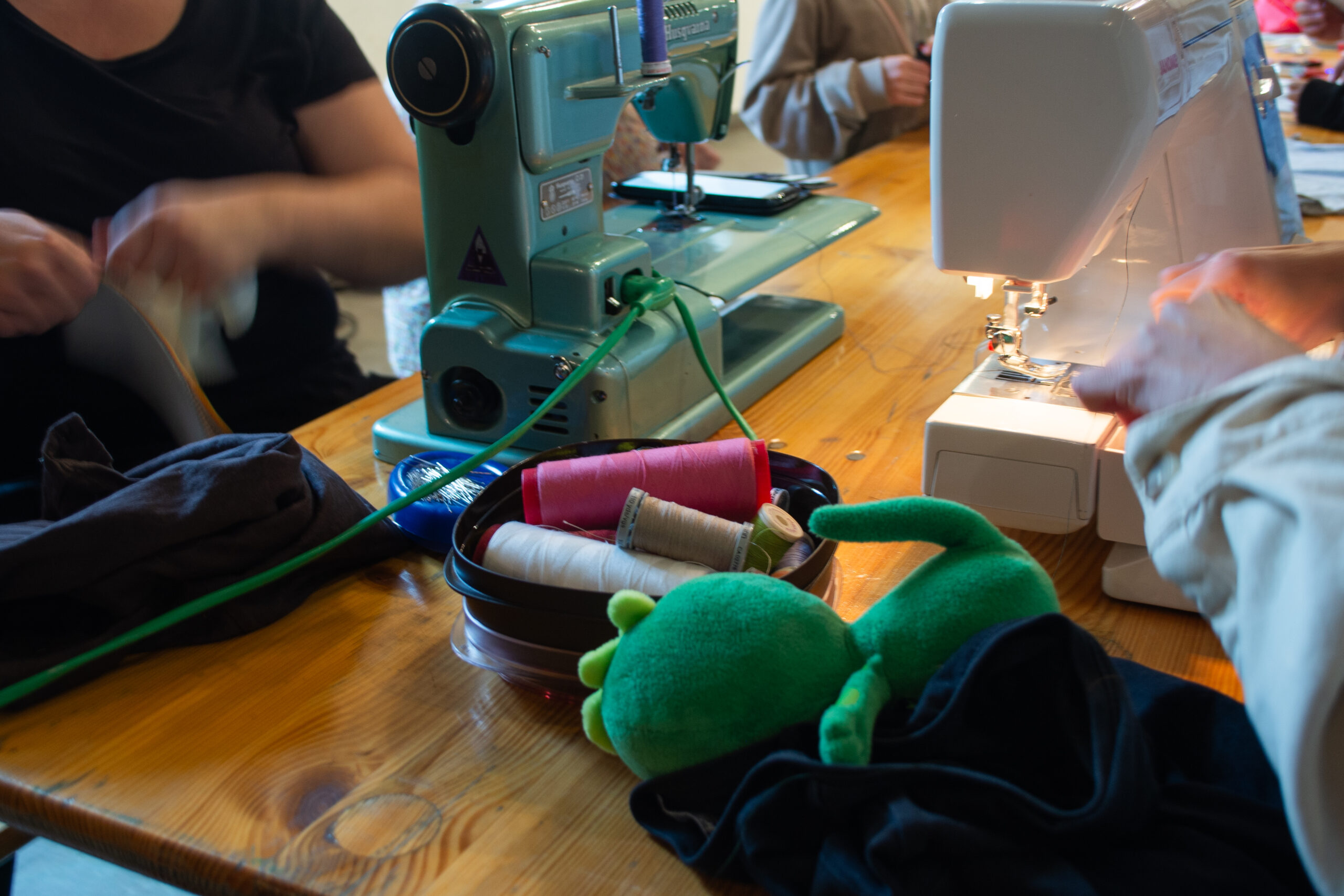 Närbild på två symaskiner och symaterial på ett bord. Två personer använder symaskinerna. 