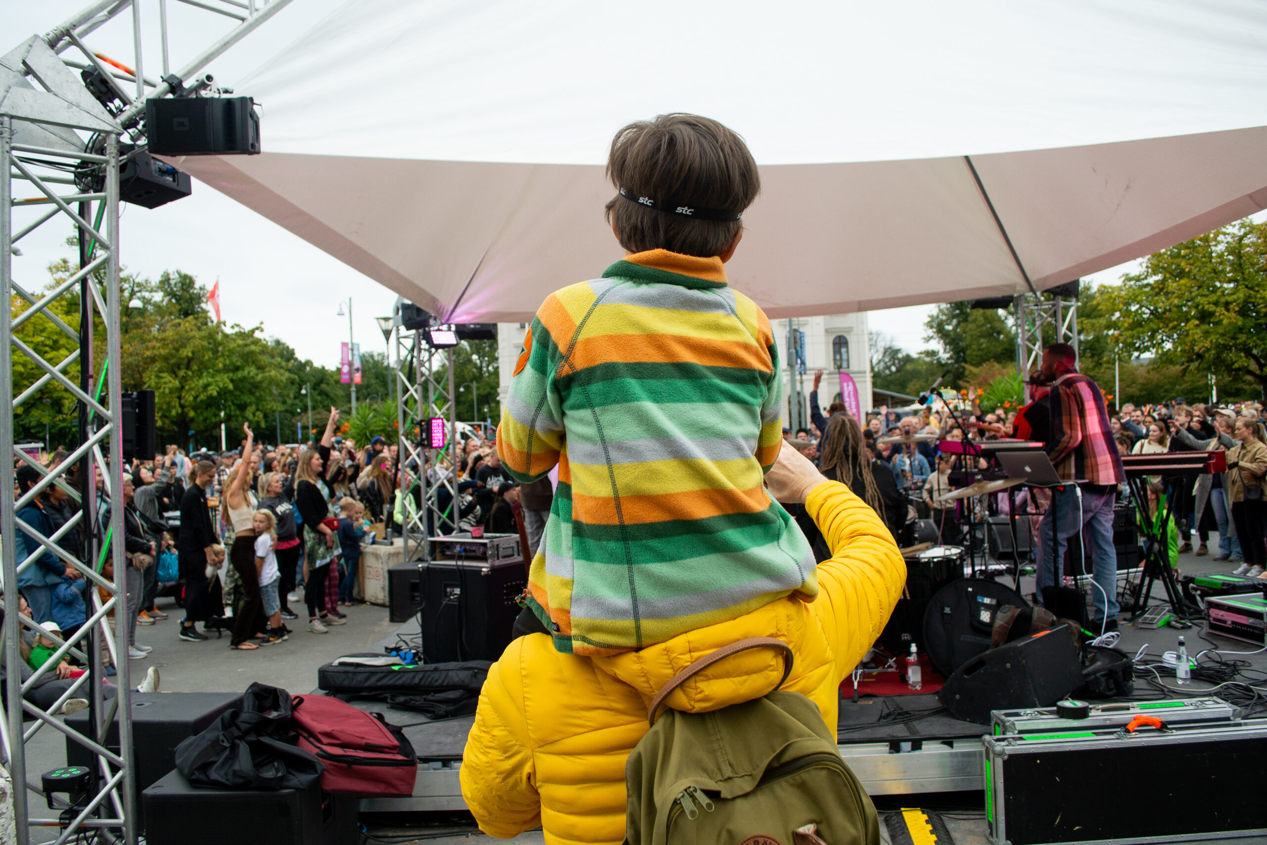A child sits on an adult's shoulders and watches a concert in Bältespännarparken. Their backs are facing the camera. The child is wearing a shirt with yellow, green and white stripes and his hair is dark brown. The adult wears a yellow sweater and moss green backpack.