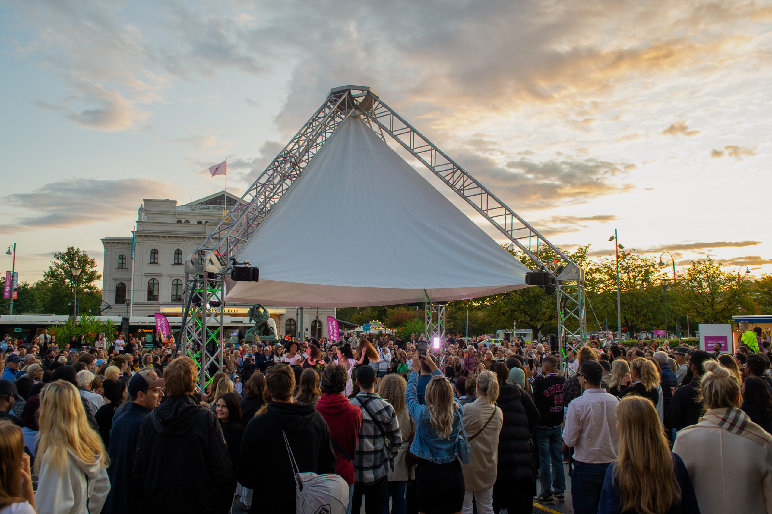 A smaller stage with a large white roof in Bältespännarparken. People perform on stage and the audience stands around the stage. The photo was taken at sunset. In the background you can see the Stora Teatern and a tram passing by.